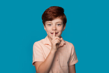 Ginger caucasian boy is gesturing the silence soung smiling on a blue studio wall in casual clothes