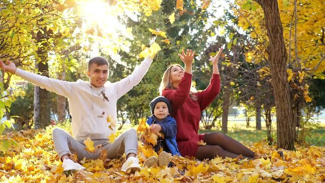 Lovely young family of mother father and cute little boy throw dry yellow leaves sitting on large pile in sunny autumn park slow motion