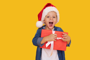 Portrait of a satisfied little child boy in christmas Santa hat. laughing isolated over yellow background. Holds a gift box. Preparing for the New Year holidays