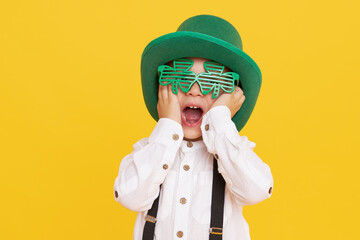 Smiling little child boy in green leprechaun hat on yellow background. St. Patrick Day celebration....