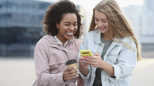 Young women friends using smartphone in a city - Cute smiling girls having fun using mobile phone on street - People, technology, friendship, relationship, mobile app, lifestyle, social media concept