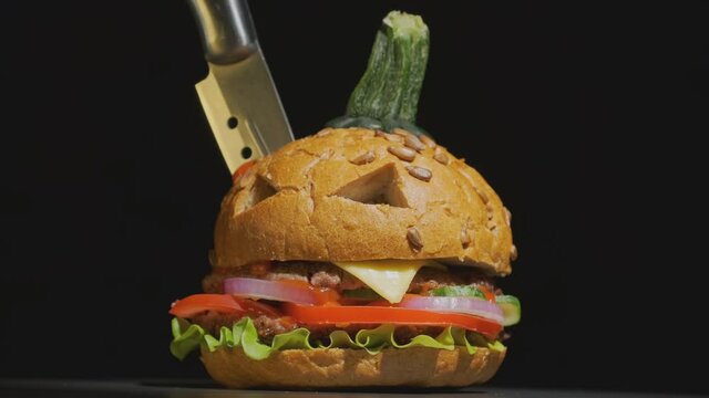 Halloween beef burger in image of pumpkin decorated with knife rotating on table on black background.