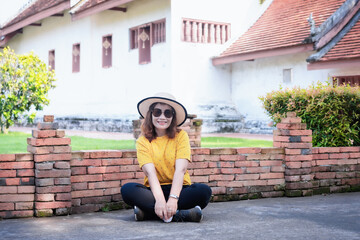 Portrait of adult beautiful Asian woman wearing a straw hat and sunglasses sitting on the floor with a temple background. Happy lady smiling and relaxing on summer vacation.