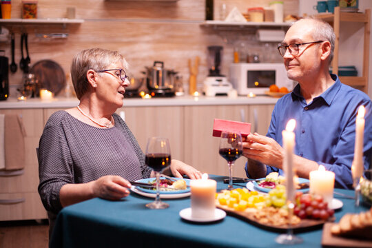 Senior Man Surprising Wife With Gift Box During Dinner. Happy Cheerful Elderly Couple Dining Together At Home, Enjoying The Meal, Celebrating Their Anniversary, Surprise Holiday