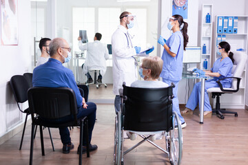Medical team with face mask against coronavirus discussing about patient treatment in hospital lobby. Assistant working on reception computer. Disabled senior woman.