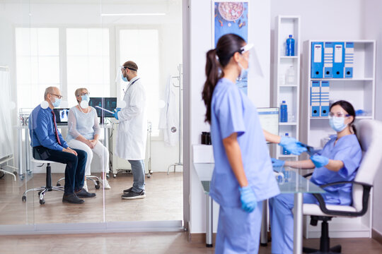 Medical Staff Wearing Face Mask Against Coronavirus In Hospital Hallway And Doctor Having A Discussion With Old Couple In Examination Room. Assistant Working On Reception Computer.