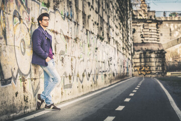 Handsome and charming young man outdoors leans against a wall in the city. Dressed in fashionable clothes and wearing eyeglasses. Coat and jeans.