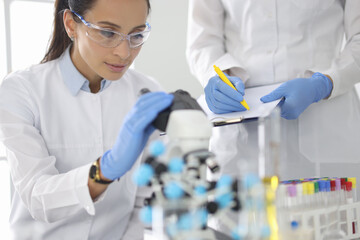 Young woman in white uniform wearing protective chemical goggles and rubber gloves looks into microscope. Biological research concept.