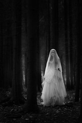 Black and white portrait of a ghost bride in a long white dress and veil standing in a gloomy dark forest.