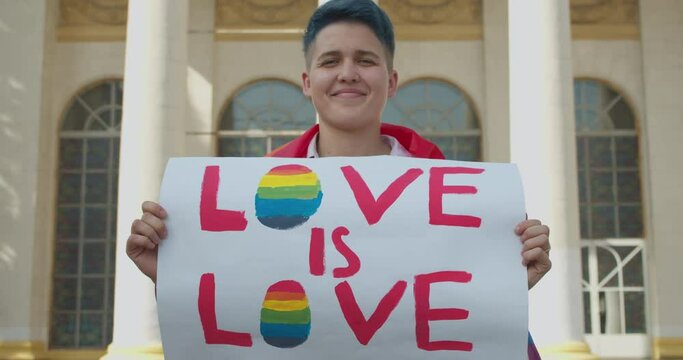 Gay girl showing love is love sign, wrapped in lgbt rainbow flag at pride parade