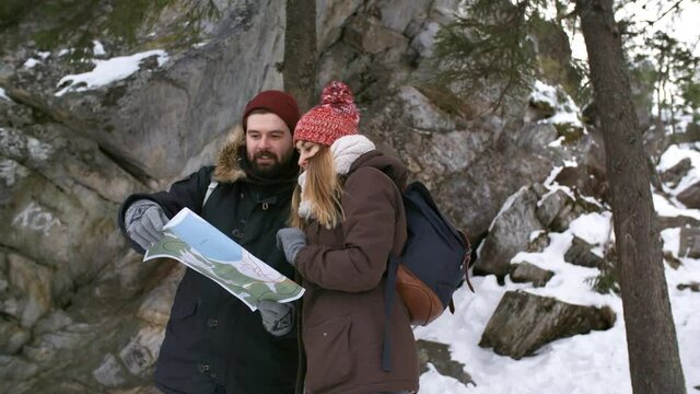 Handheld medium shot of happy young man and woman with backpacks looking at paper map and discussing the safest trail while hiking in mountains in winter