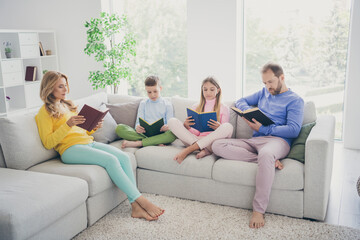 Full size photo of focused family mom dad two little kids boy girl sit couch read book in house indoors