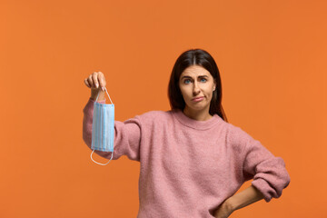 I give up. I don't care about the virus. Dissatisfied young female holds medical mask. looks sadly at camera. does not want to use it. wears in pink knitted sweater isolated over orange wall