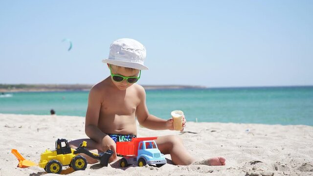 Sunny summer day, a boy plays with sand and toys, cars on the beach. a child eats ice cream while sitting on a sandy beach with an azure sea background.