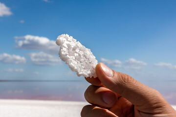 Hand fingers holding white shining salt flake crystal with blurred natural pink salty lake and blue sky background. Spa resort sunny close-up on Syvash, Ukraine
