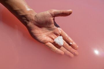 Hand holding white salt crystal in pink vibrant salty water sunny close-up. Spa procedures in...