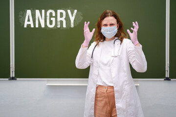 Angry school nurse in a medical mask on a blackboard background, copy space. Concept of problems at...