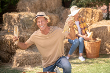 Happy male farmer showing cobs into the camera, female farmer shucking cobs behind him