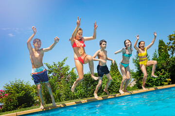 Group of kids jump with lifted hands in water pool outside during summer vacation side view