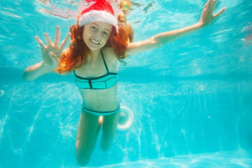 Obraz na płótnie Canvas Happy girl dive and swim underwater wearing Santa Claus hat in the pool smile on her New Year vacation