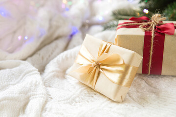 A box with a Christmas gift in light paper is tied with a gold ribbon on a white knitted background with decor. Christmas tree pendant, green branches, lights garlands. New Year. space for text