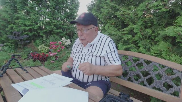 Pensioner with glasses turns over pages and marks text in instruction of digital camera sitting at table in cottage house garden
