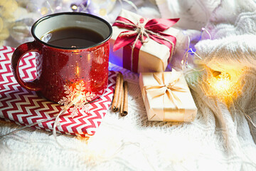 Red mug with tea in Christmas decorations on a white knitted blanket. New year, garlands, gifts, cinnamon and dried orange - an atmosphere of warmth, comfort and magic.