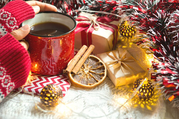 Naklejka na ściany i meble A woman's hand in a warm sweater holds a red mug with a hot drink on a table with Christmas decorations. New year's atmosphere, cinnamon sticks and a slice of dried orange, gifts, garland and tinsel