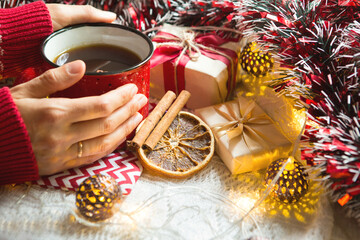 A woman's hand in a warm sweater holds a red mug with a hot drink on a table with Christmas decorations. New year's atmosphere, cinnamon sticks and a slice of dried orange, gifts, garland and tinsel