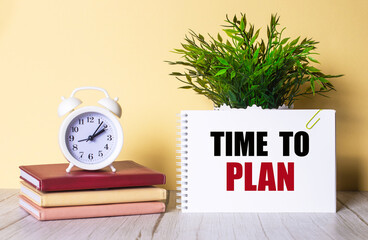 TIME TO PLAN is written in a notebook next to a green plant and a white alarm clock, which stands...