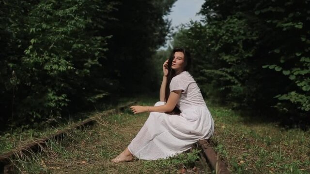 A romantic young girl in a white dress sits barefoot on the rails of an old abandoned railway against the backdrop of green trees. Side view. Close-up. Slow motion