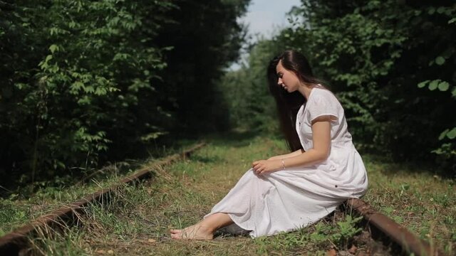 A modest young girl sits barefoot on the tracks of an old abandoned railroad in the woods and tilting her head straightening her long white dress. Side view. Close-up