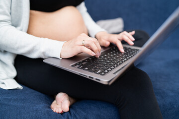 A pregnant woman wearing sports clothes sits on the couch in the living room and texting an email to a specialist doctor on a laptop.