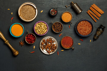 Bright spices and fragrant dried herbs in colored bowls, pepper mill on a black background. Top...