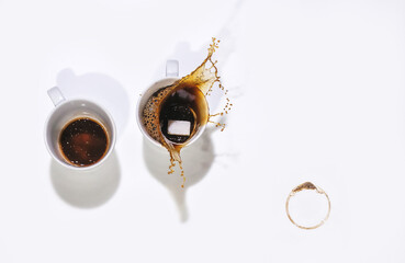 A sugar cube falls into a white Cup of coffee, splashing. Another empty cup of coffe and a coffee cup stain. Top view, white background and copy space