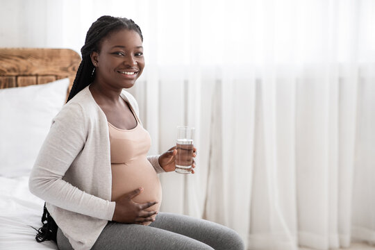 Happy African Pregnant Woman Drinking Water From Glass While Relaxing On Bed