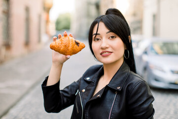Close up face portrait of young charming smiling Asian lady in black leather jacket, posing to camera outdoor in old European city, holding fresh croissant. People, food, urban lifestyle