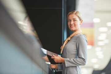 Waist up portrait of elegant flight attendant standing by check in desk and smiling at camera holding tickets, copy space