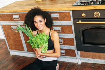 A cheerful african american woman in sportswear with an armful of fresh vegetables and greenery in...