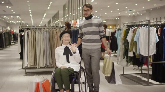 Portrait shot with tracking of happy paraplegic woman in wheelchair and her male friend holding shopping bags chatting in clothing store, then looking at camera and smiling