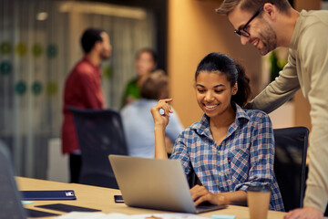 Young cheerful caucasian man helping female coworker with computer work, two young multiracial colleagues using laptop and discussing project