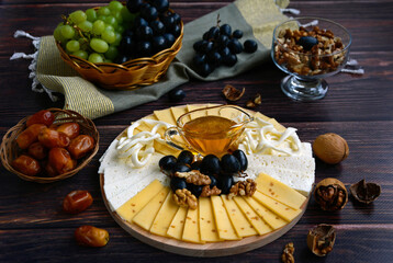 Different types of cheese on a wooden plate, green and black grapes, walnut, dates, napkin are located on a dark wooden table