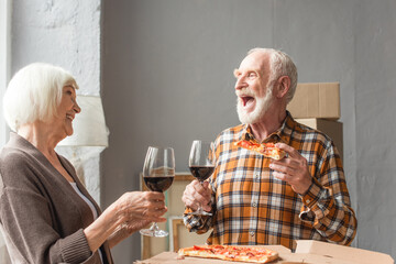 laughing senior man holding piece of pizza and looking at wife while holding glasses of wine