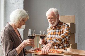 senior couple holding glasses of wine and man taking piece of pizza in new house