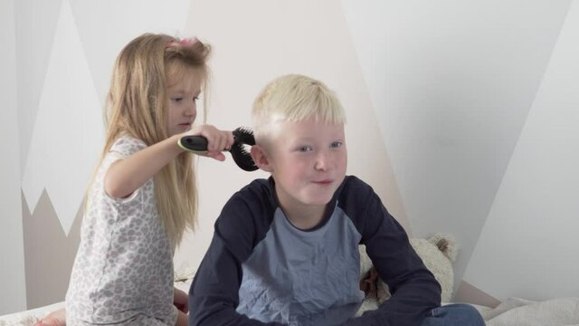 Funny Little Sister combs blond hair to her older brother. Children play in a beauty salon.