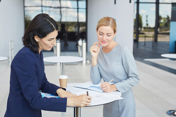 Fototapeta premium Waist up portrait of two successful businesswomen discussing deal while standing by cafe table in airport or office building