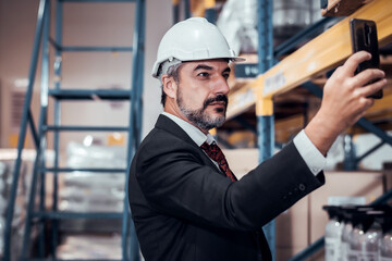 Businessman wearing a hardhat standing at goods warehouse and check for control loading from Cargo freight ship for import and export by report by smartphone. commercial warehouse for trading concept 