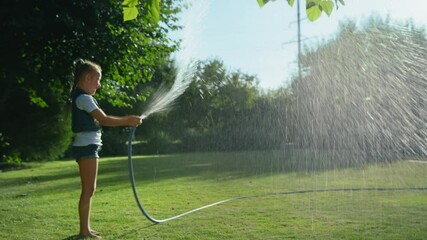 adorable little girl playing with a garden hose on hot and sunny summer evening, child watering green grass