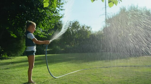 adorable little girl playing with a garden hose on hot and sunny summer evening, child watering green grass