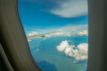 View from inside plane through plane window over blue sky, white clouds and green mountain. Commercial airline flight for travel in Thailand. Plane wing above the city. Airplane flying on blue sky.